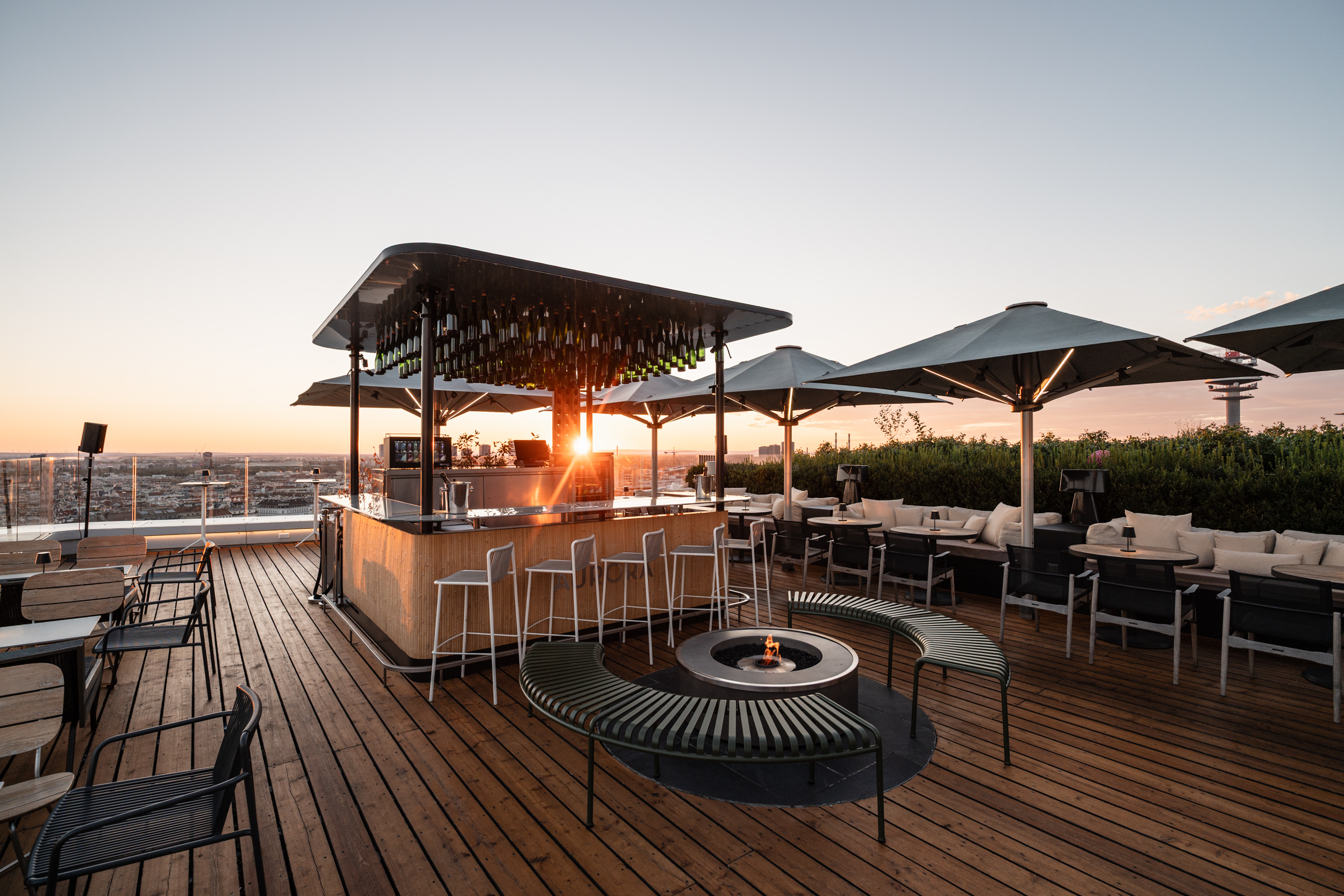 The Aurora rooftop bar at sunset: the bar counter, a fire pit and seating on a timber deck, with Vienna beyond